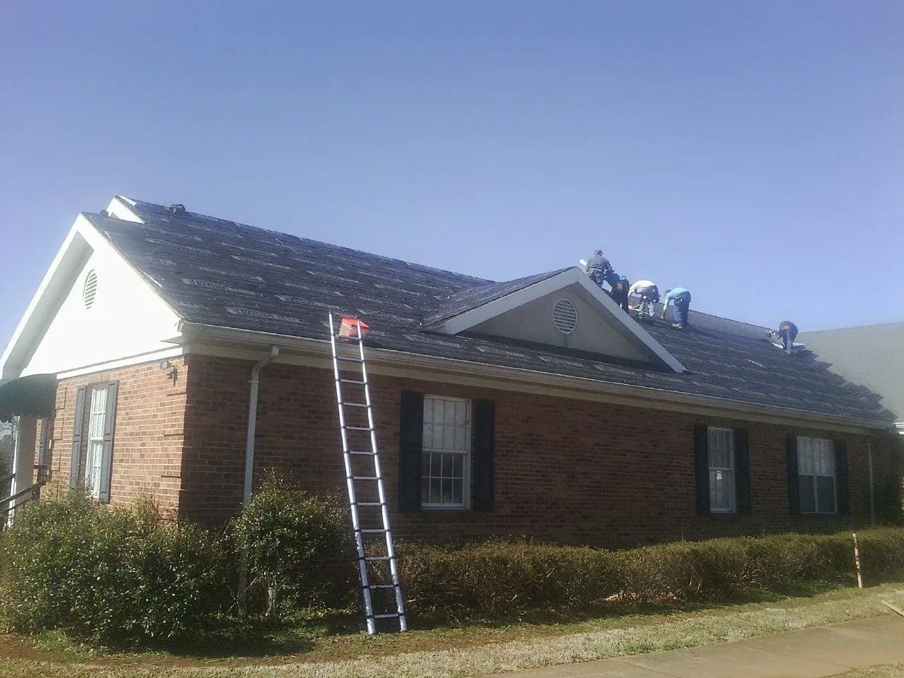 Crew tearing shingles off a brick ranch roof with a ladder set — Metal Roof Repair work by our crew in Jansen