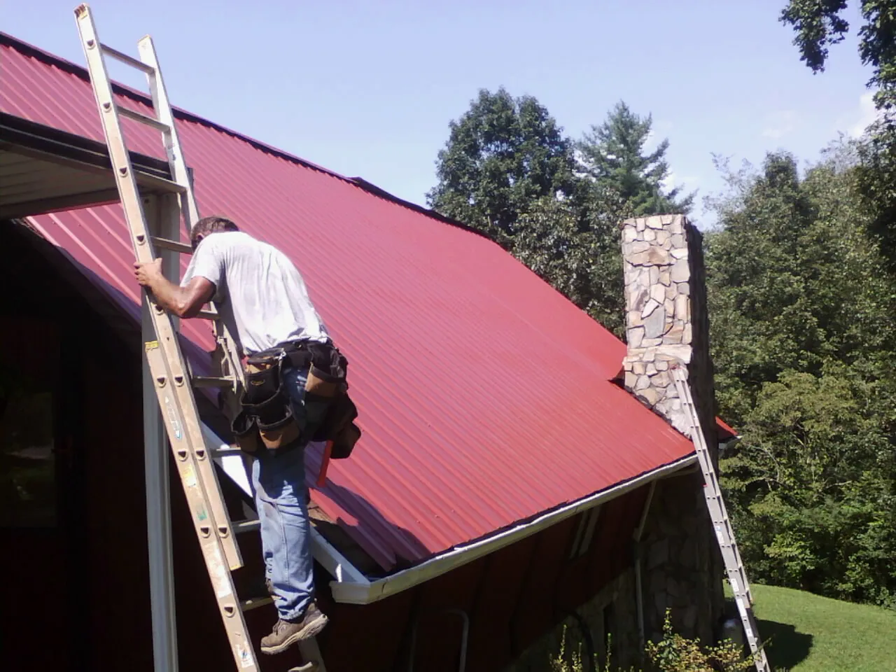 Roofer on a ladder beside a red metal roof and stone chimney — Soffit & Fascia Repair by Roofing Brothers in Jansen